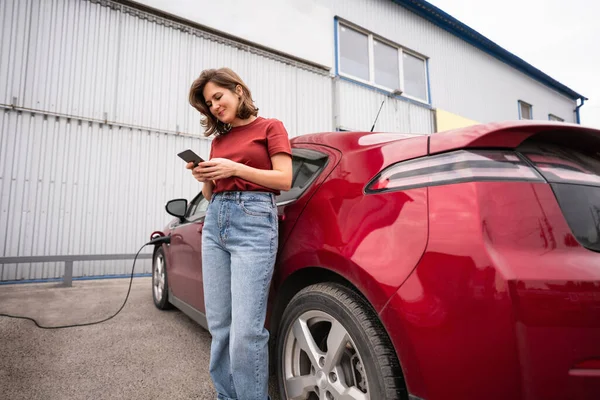 Kobieta Smartfonem Ładującym Czerwony Samochód Elektryczny — Zdjęcie stockowe