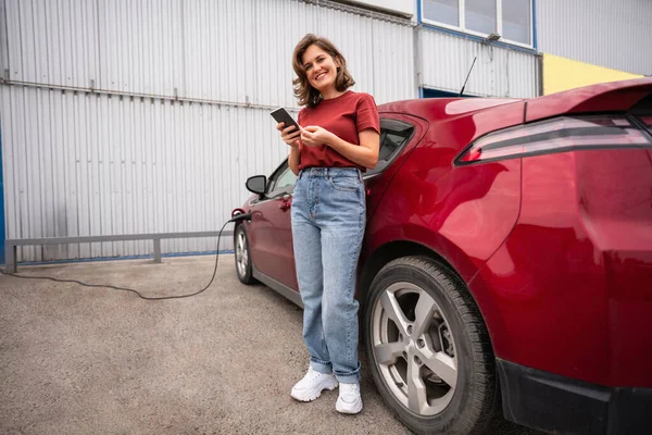 Kobieta Smartfonem Ładującym Czerwony Samochód Elektryczny — Zdjęcie stockowe