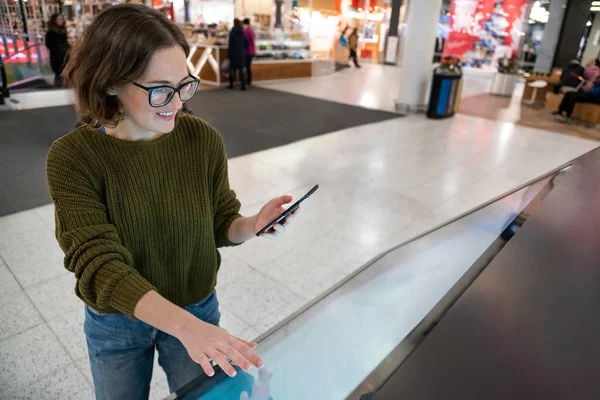Woman with phone uses self-service kiosk in the shopping mall.
