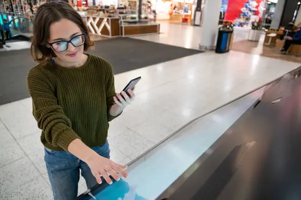 Woman with phone uses self-service kiosk in the shopping mall.