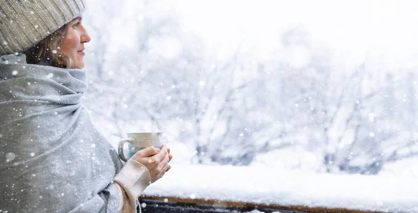 暖かい帽子と毛布の女性は 彼女の手に熱い飲み物を持ったマグカップを持っています 背景に雪が降る冬の風景 — ストック写真