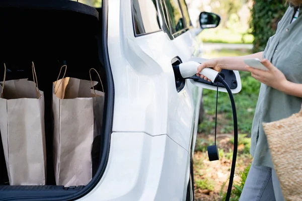 カントリーハウスの庭の充電電電気自動車の隣にショッピングバッグを持つ女性 — ストック写真