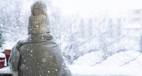 テラスの暖かい帽子と毛布の女性 背景に雪が降る冬の都市風景 — ストック写真