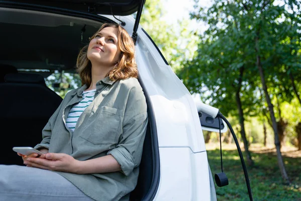 スマートフォンを持つ女性は電気自動車のトランクに座ります — ストック写真