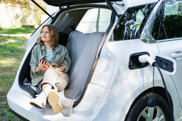 Akıllı Telefonlu Bir Kadın Elektrikli Bir Arabanın Bagajında Oturuyor — Stok fotoğraf