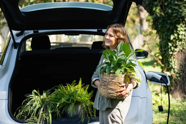 カントリーハウスの庭の充電電電気自動車の隣に鍋に植物を持っている女性 — ストック写真