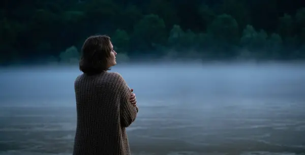 A woman in a sweater on the bank of a foggy river in the evening,