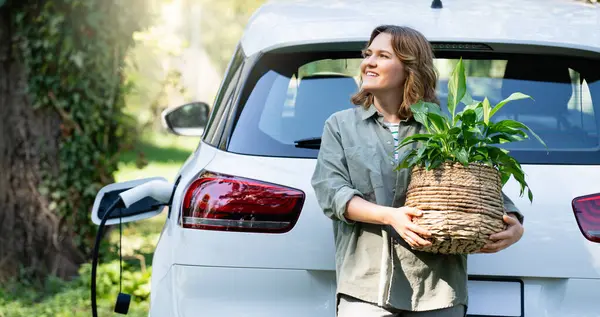 カントリーハウスの庭の充電電電気自動車の隣に鍋に植物を持っている女性 — ストック写真