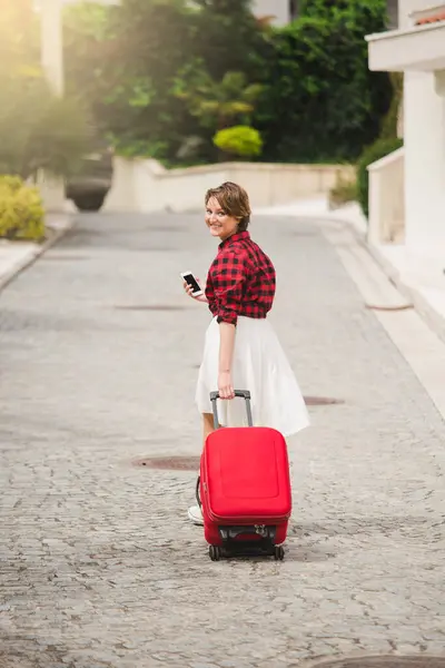 Young woman in a red shirt with a suitcase and phone on the city street.