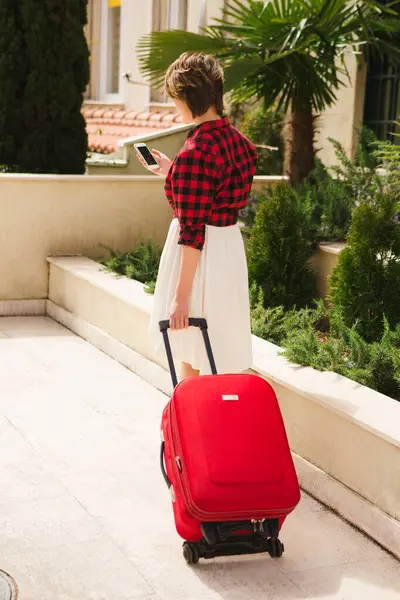 Young woman in a red shirt with a suitcase and phone on the city street.