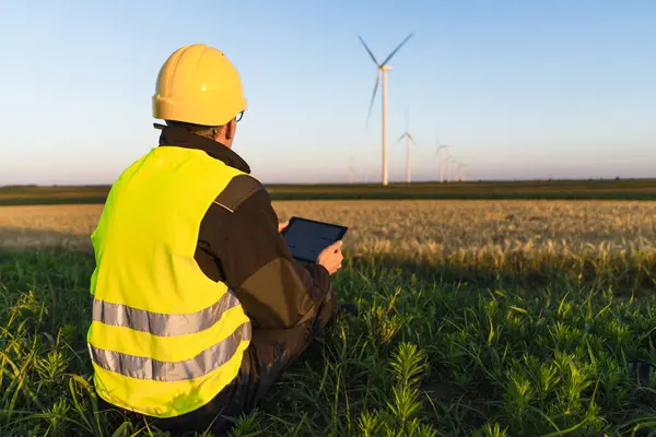 Stock image Worker with digital tablet sits on the grass and looks at wind turbines..