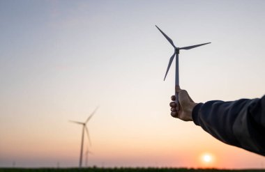 Man holds toy wind turbine in his hand. Real wind turbines in the background. clipart