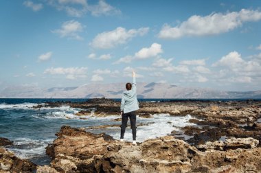 Akdeniz 'in kayalık sahillerinde güzel bir kız rahatlar ve elini kaldırır. Akdeniz ve Girit 'in Rocky Sahili, Yunanistan Yeri Balos yakınlarında.
