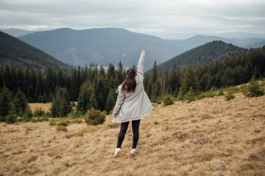 Happy girl stands in the mountains, enjoys the beautiful view and raises her hand up.