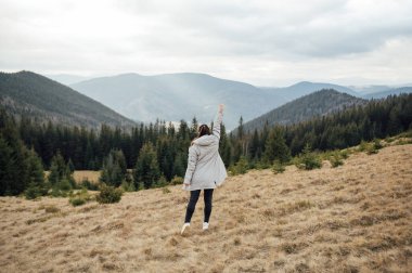Happy girl stands in the mountains, enjoys the beautiful view and raises her hand up.