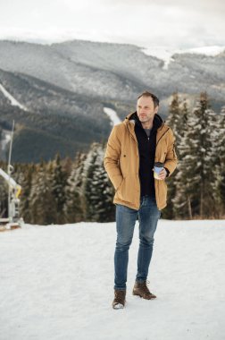 Morning coffee in the mountains. Handsome man drinking coffee and looking at the mountains in Ukraine. Snowy mountain background. Winter holidays, tourism, travel and people concept.