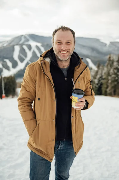 Morning coffee in the mountains. Handsome man drinking coffee and looking at the mountains in Ukraine. Snowy mountain background. Winter holidays, tourism, travel and people concept.