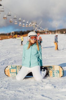 A beautiful girl snowboarder sits on a slope, resting after a difficult descent. Sunny day