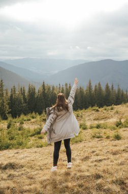 Happy girl stands in the mountains, enjoys the beautiful view and raises her hand up.