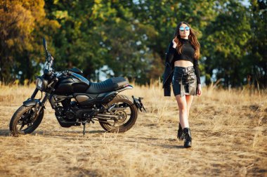 Siyah elbiseli güzel bir motosikletli kız elinde kaskla motosikletin başında duruyor..