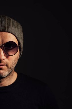 Güneş gözlüklü ciddi bir dramatik adam siyah stüdyonun arka planında dikilirken başka tarafa bakıyor..