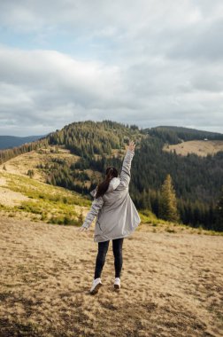 Happy girl stands in the mountains, enjoys the beautiful view and raises her hand up.