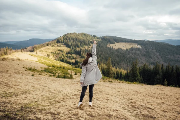 stock image Happy girl stands in the mountains, enjoys the beautiful view and raises her hand up.