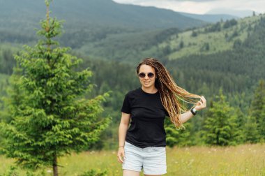 Saç stili olan güzel bir kadın. Bir sürü örgülü. Siyah tişört ve şortla dağın tepesinde poz veriyor.