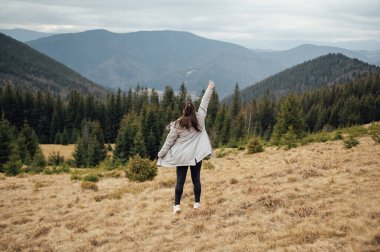 Happy girl stands in the mountains, enjoys the beautiful view and raises her hand up.