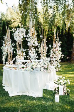 Yeni evliler için düğün masası. Beyaz tabaklar ve peçetelerle süslenmiş şölen masası, yeşillik ve çiçekler ve mumlar, açık havada.