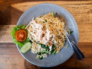 Mie Goreng Jawa, geleneksel kızarmış Java eriştesi dilimlenmiş tavuk, marul, salatalık ve domates ile ahşap bir masanın üzerinde gri bir tabakta servis ediliyor.