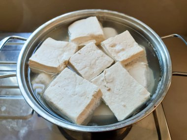Taze tofu kalıplarının paslanmaz çelik bir tencerede pişirilmeye hazır halde suya batırılmış yakın plan görüntüsü. Mutfak, sağlıklı beslenme ve vejetaryen içeriği için ideal.