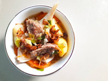 Kızarmış arpacık soğanları ve yeşil soğanlarla süslenmiş kemikli et, patates ve havuçla bembeyaz bir kasede servis edilen et çorbası.