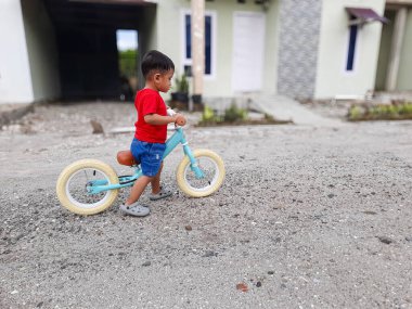 Kırmızı tişörtlü ve kot şortlu bir çocuk modern bir evin önündeki çakıllı bir yolda turkuaz denge bisikleti sürmeyi öğreniyor. Çocukluk, açık hava ve aile temaları için ideal