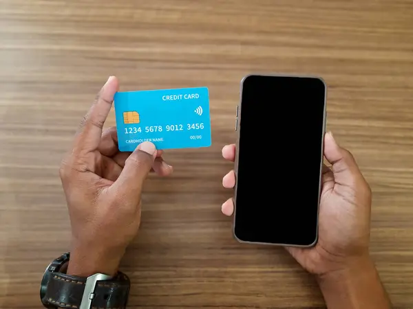 Adamın biri ahşap bir masada mavi bir kredi kartı ve akıllı telefon tutuyor. Fintech işlemlerini ve online ödemeleri gösteriyor.