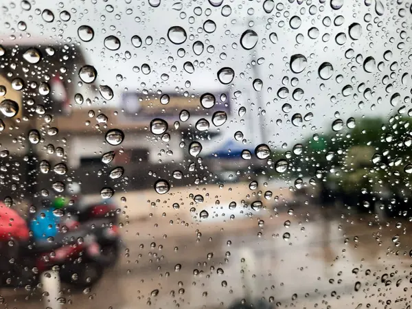 Yağmurlu bir günde cam bir penceredeki su damlacıklarının yakın görüntüsü arka planda bulanık bir şehir manzarası ve sakin, karamsar bir atmosfer yaratıyor.