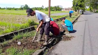 Pariaman, Endonezya - 11 Haziran 2025: Küba Hava Köyü 'nde yapılan bir temizlik etkinliği sırasında geleneksel aletlerle yol kenarı çimlerini budayarak erkek işçiler