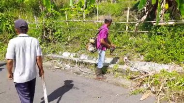 Pariaman, Endonezya - 25 Haziran 2025: Bir erkek işçi Sungai Rambai köyündeki bir temizlik etkinliği sırasında yol kenarındaki çim biçme makinesini kullanarak çim biçiyor