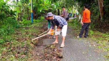 Pariaman, Endonezya - 9 Temmuz 2025: ortak bir çalışma sırasında çapa kullanarak asfalt bir yolun kenarını temizleyen adamlar. Bataklık köyündeki yol kenarını kaplayan toprak ve otlar temizleniyor.