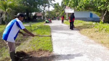 Pariaman, Endonezya - 23 Haziran 2025: bir adam yolu genişletmek için çapa kullanarak asfalt yolun kenarını temizler. Manggung köyündeki yol kenarını kaplayan toprak ve otlar temizleniyor.