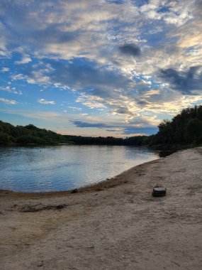 Kumlu sakin bir nehir, gün batımında gökyüzünü ve bulutları yansıtan su.