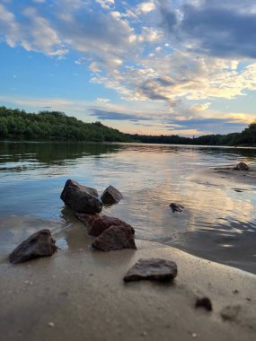 Sudaki büyük kayalar ve gökyüzündeki ılık tonlar sakin bir nehrin üzerinde canlı bir günbatımı manzarası yaratır.