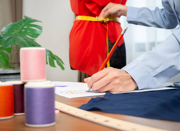 Genç Asyalı kadın moda tasarımcısı manken üzerinde ölçü alıyor ve stüdyosundaki kağıda yeni bir tasarım çiziyor.