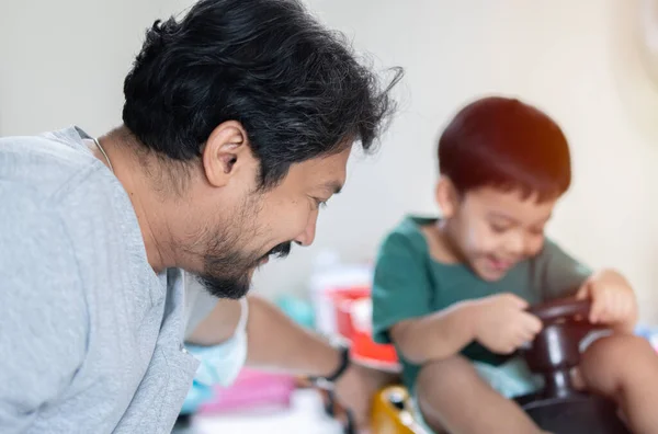 Asian dad sitting on floor playing with toddler son with toy cars at home, happy caring black father have fun with little kid engaged in funny activity race on automobiles.