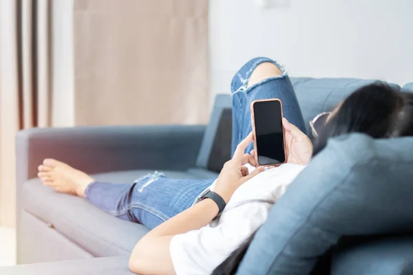 Asian woman enjoys online communication while lying on blue couch in living room. Technology, communication concept