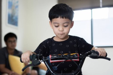 Çocuk kasıtlı olarak evde bisiklet sürmeyi öğreniyor, babası yakınlarda izlerken. Mutlu bir çocukluk kavramı.