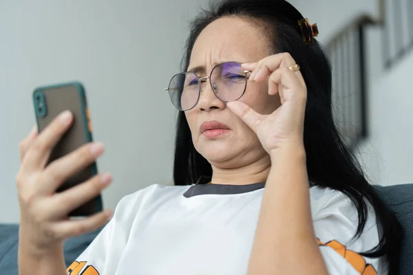 Asian senior fatigue woman try to move eye glasses during using smartphone after surfing internet or social media at home. problem or blurry vision from old aged concept.