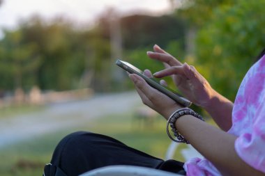 Asyalı kadın elleri, bulanık yeşil park arka planına karşı akıllı telefon tutuyor. Yakın plan, boşluğu kopyala