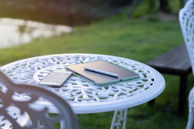 Akıtılmış akıllı telefon ve tablet manzaralı yeşil çimenlikteki beyaz bahçe masasında. Yan görüş. Doğal arkaplan.