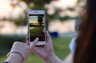 Genç bayan hipster, mobil cihazlar için fotoğraf uygulaması aracılığıyla internet sosyal medyasında paylaşmak için akıllı telefon kamerasıyla nehir manzarasının fotoğrafını çekiyor..